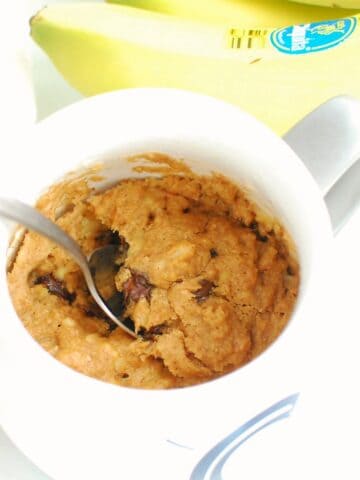 A spoon digging into a healthy banana mug cake, next to some fresh bananas and chocolate chips.