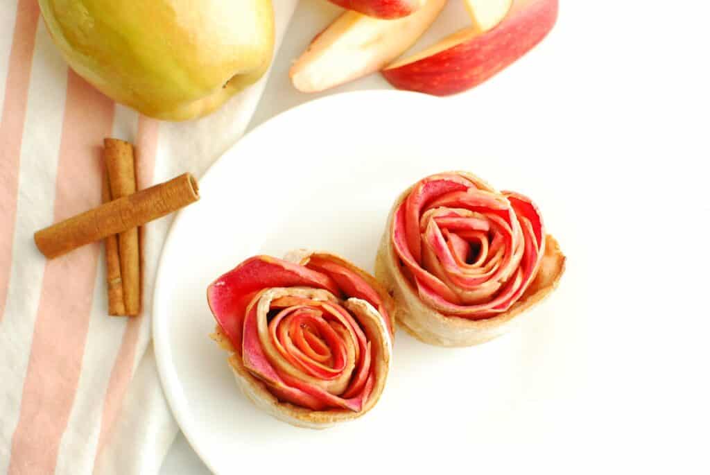 Two apple roses on a plate next to cinnamon sticks and fresh apples and a napkin.