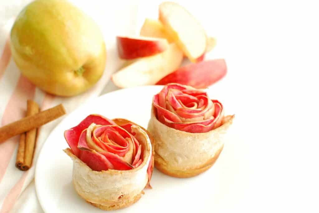 Two baked apple roses on a plate with a whole apple and sliced apple in the background.