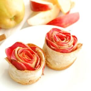 Two apple roses on a plate with fresh sliced apples in the background.