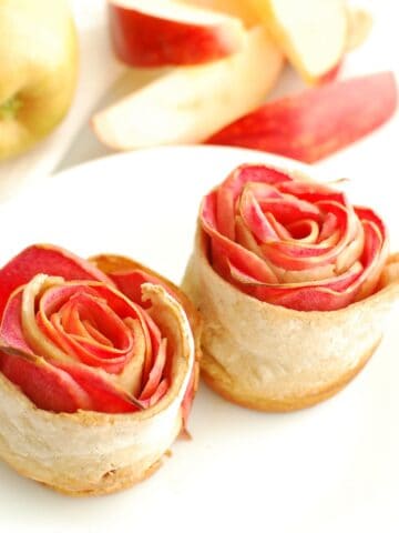 Two apple roses on a plate with fresh sliced apples in the background.