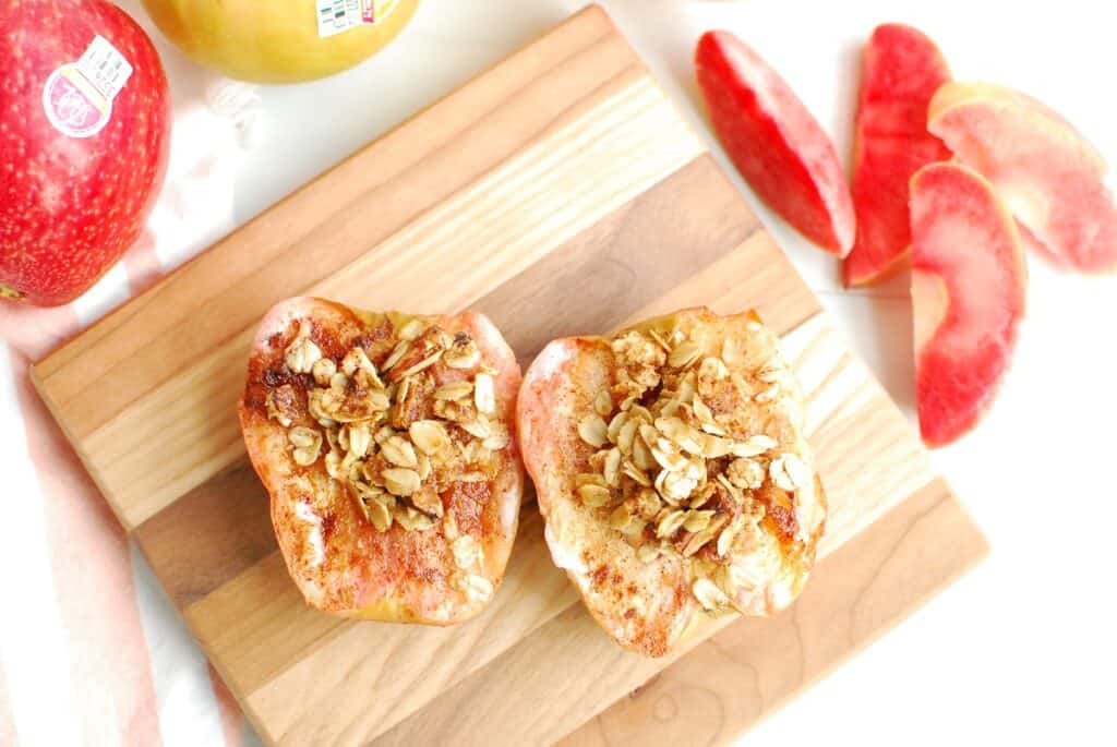 Air fryer baked apples on a small wooden cutting board next to fresh apple slices and whole apples.