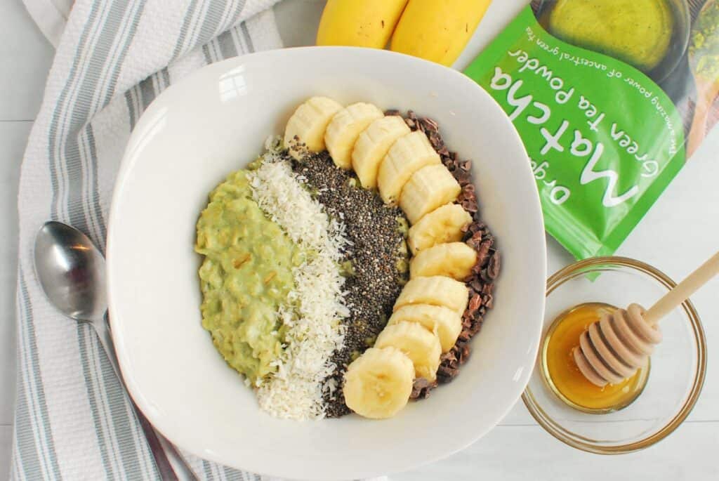 A bowl of matcha oatmeal with toppings next to a package of matcha powder and a bowl with honey.