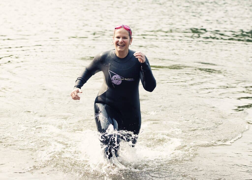 A woman running out of a lake in a wetsuit after having gone for a swim.