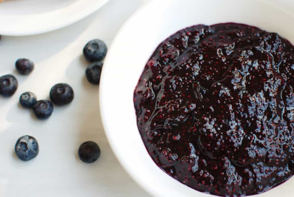 A bowl of blueberry chia jam next to some scattered fresh blueberries.