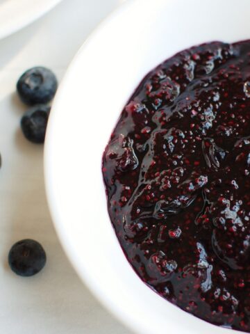 A bowl of blueberry chia jam next to some scattered fresh blueberries.