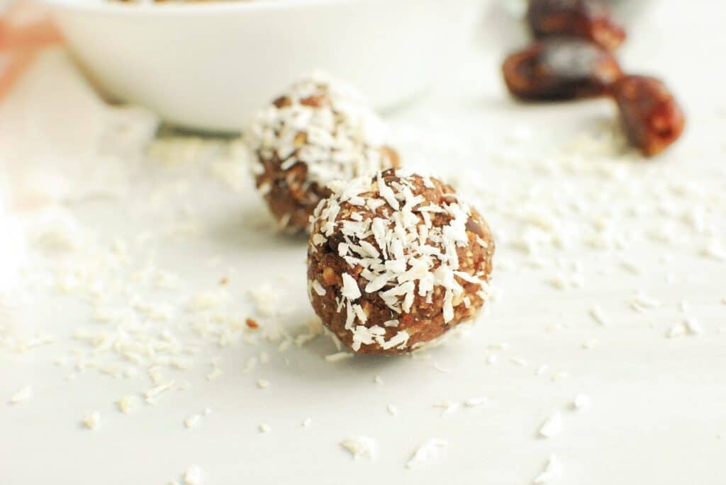 Two almond date coconut balls on a white table with scattered shredded coconut around them.