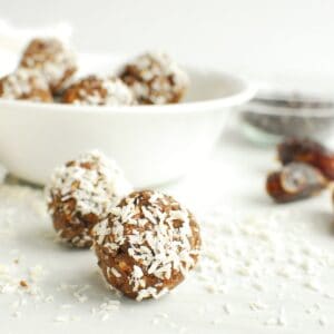 Two coconut almond date balls on a table next to shredded coconut, dates, and a bowl.