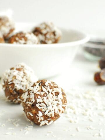 Two coconut almond date balls on a table next to shredded coconut, dates, and a bowl.