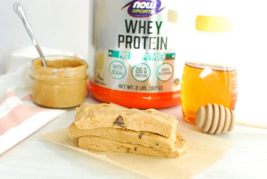 A stack of three peanut butter protein bars next to a container of protein powder, honey, and peanut butter.