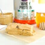 Three peanut butter protein bars next to a container of protein powder, a jar of peanut butter, and a bottle of honey.