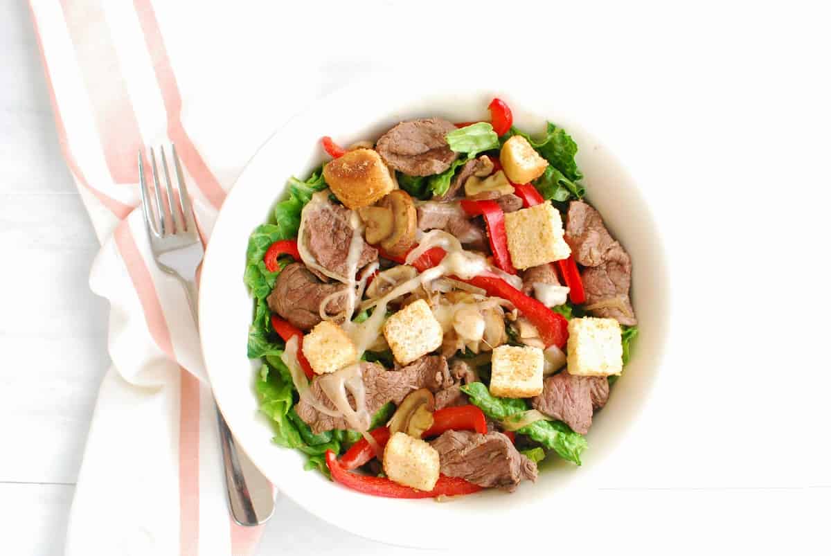 A bowl of cheesesteak salad next to a napkin and fork.