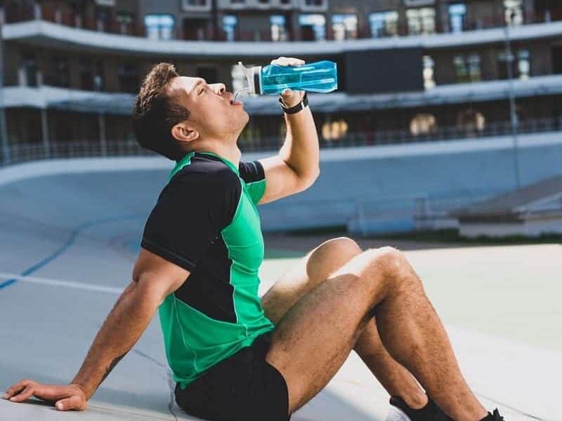 A male runner sitting at an outdoor track drinking water.