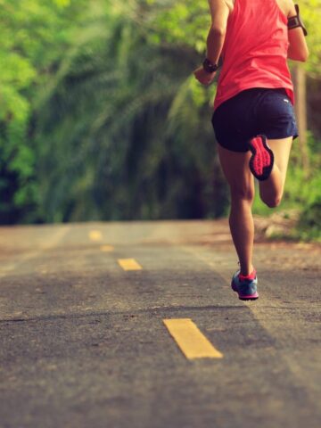 A man in a red tank top and black shorts running on the road to train for a 5K.