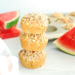 A stack of three coconut watermelon muffins with slices of watermelon and a muffin tin in the background.