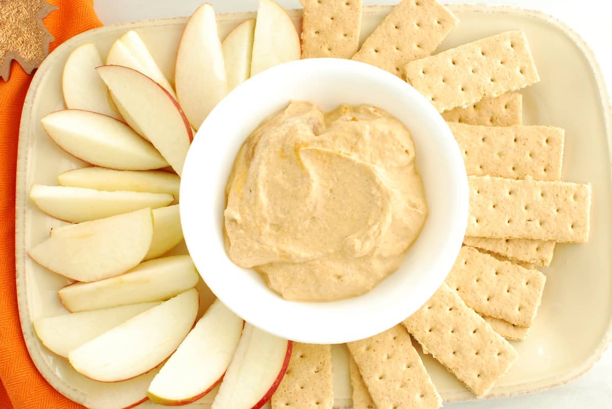 A bowl of dip on a platter surrounded by graham crackers and apple slices.