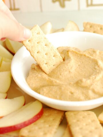 A hand dipping a graham cracker into a bowl of pumpkin greek yogurt dip.