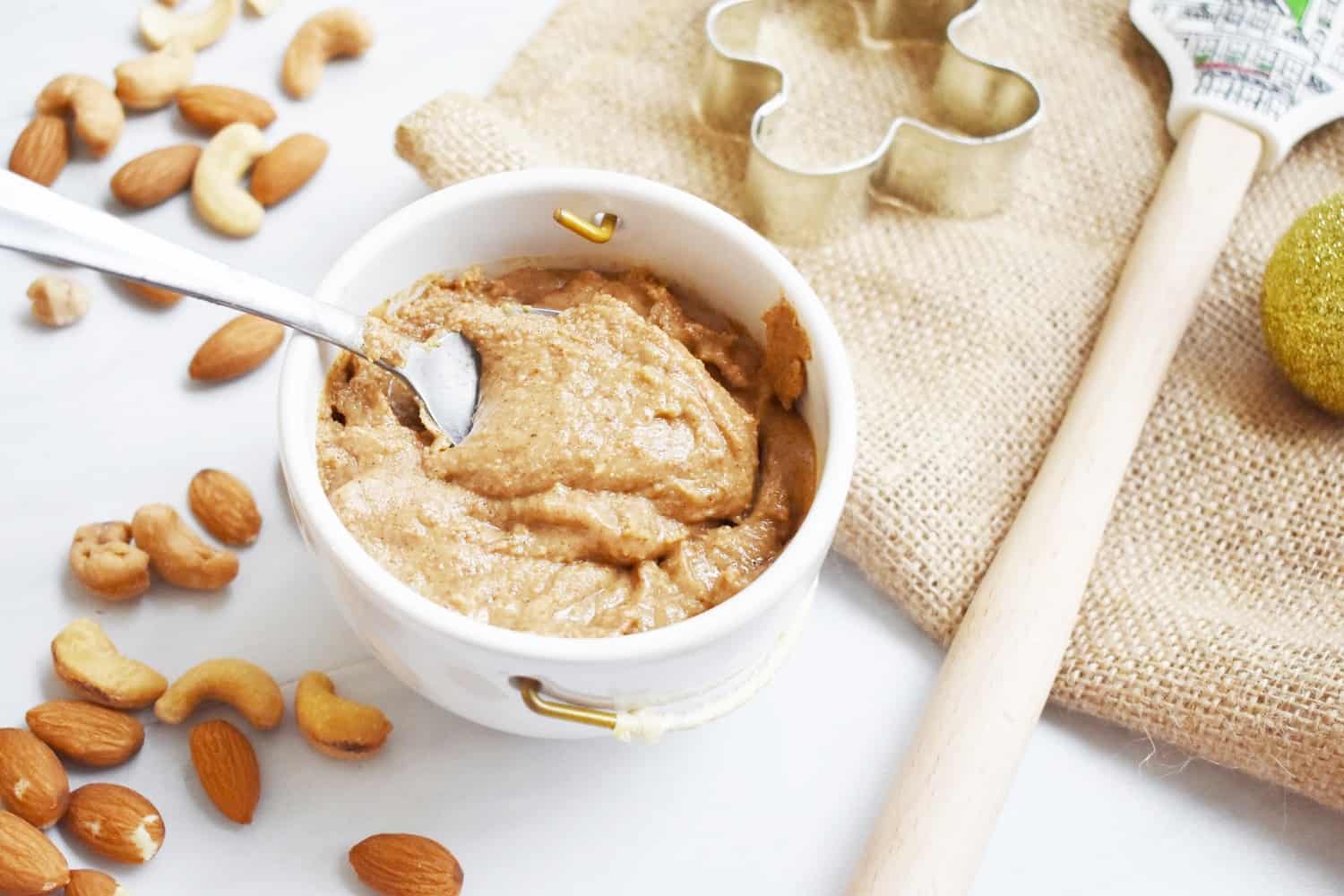 A small bowl of gingerbread nut butter with a spoon in it, next to some almonds and cashews.