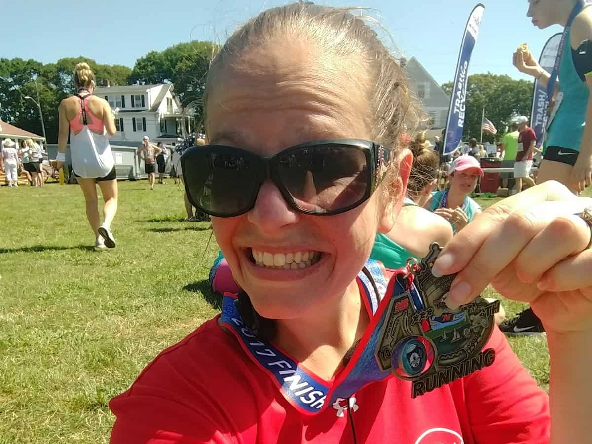 A woman holding up a medal from a road race, wearing sunglasses and a red t-shirt.