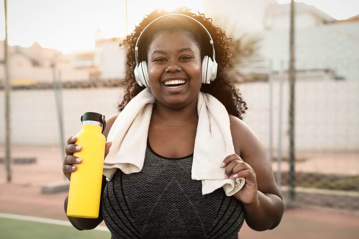 A curvy woman just finishing up a running workout, holding a water bottle and wearing headphones.