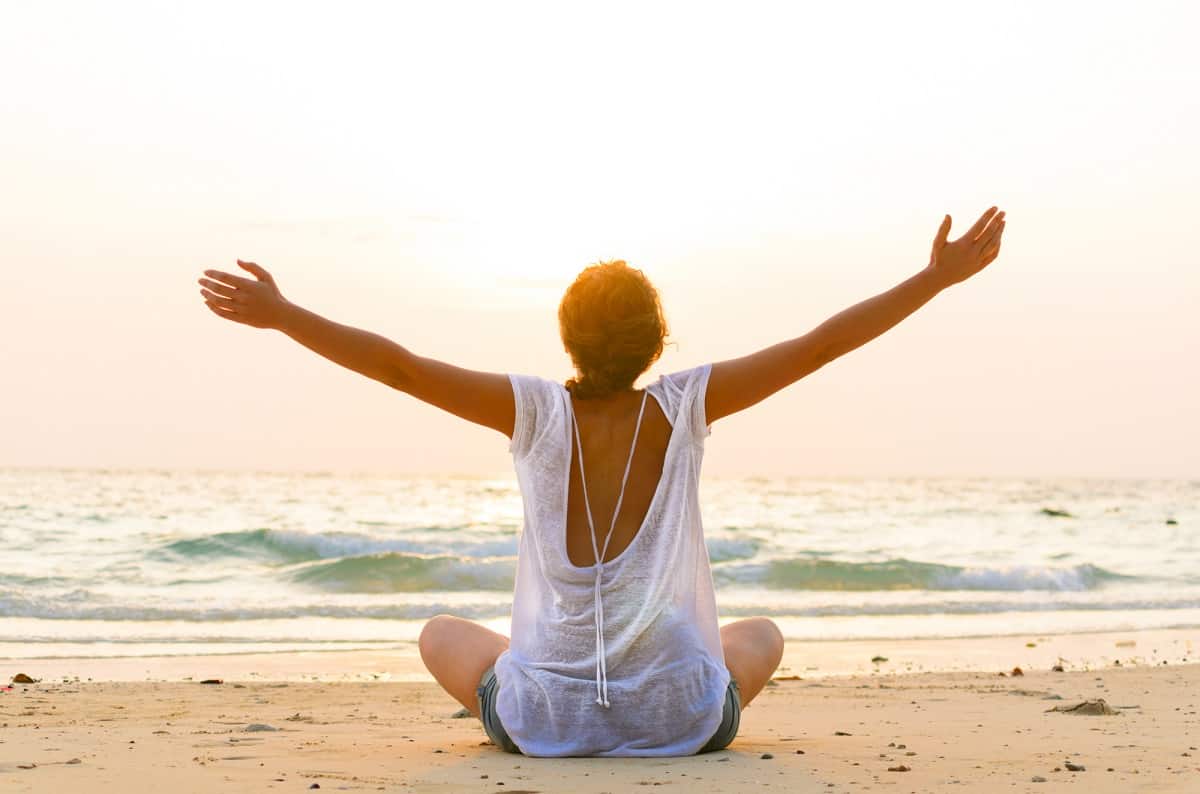 A woman sitting at the beach with her arms outstretched during summer.