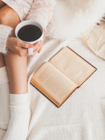 A woman reading a book while drinking a cup of coffee on a cozy bed for her winter self care activity.