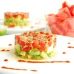 Watermelon tartare on an avocado cucumber base, on a white plate drizzled with sriracha.