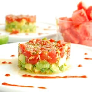 Watermelon tartare on an avocado cucumber base, on a white plate drizzled with sriracha.