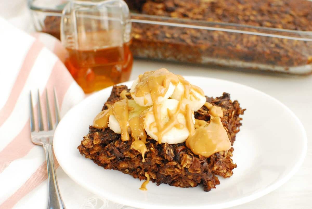 A piece of chocolate banana baked oatmeal on a white plate, topped with bananas and peanut butter.