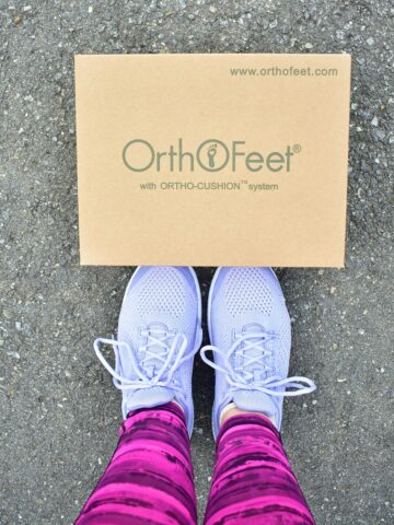 A woman's feet wearing Orthofeet sneakers standing next to a shoe box.