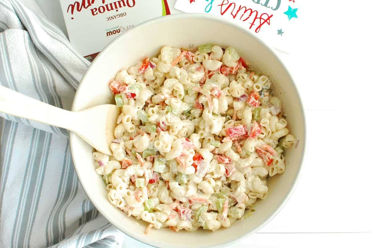 A bowl of Greek yogurt macaroni salad next to a napkin, a box of macaroni, and a summer sign.