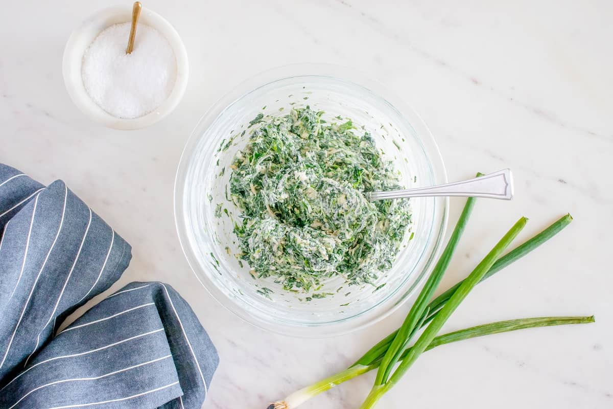 A glass mixing bowl with the spinach and cheeses mixed into the other filling ingredients.