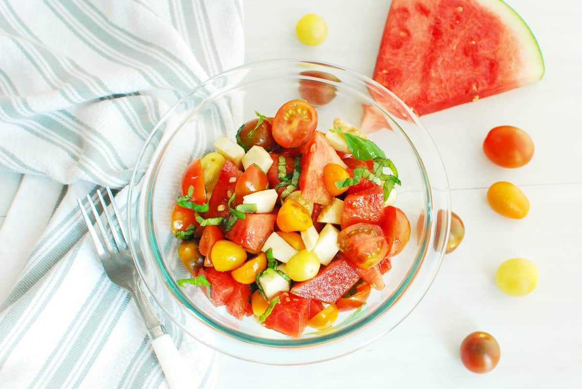 A glass bowl of watermelon caprese salad next to a napkin and fork.