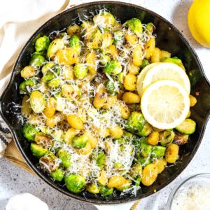 A skillet gnocchi and brussels sprouts dish, garnished with lemon, next to some fresh garlic and parmesan.