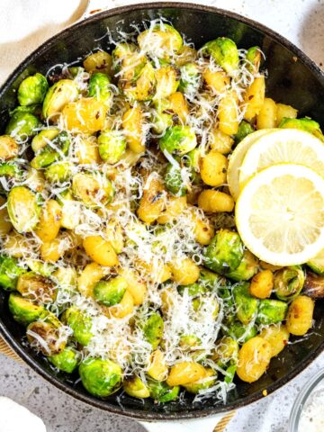 A skillet gnocchi and brussels sprouts dish, garnished with lemon, next to some fresh garlic and parmesan.