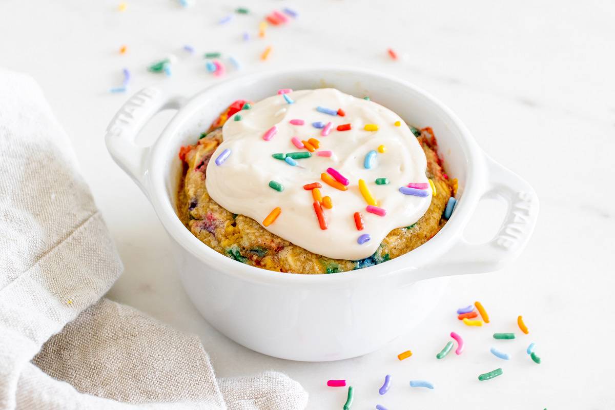 A white ceramic dish with birthday cake baked oats on a table next to a napkin and assorted sprinkles.