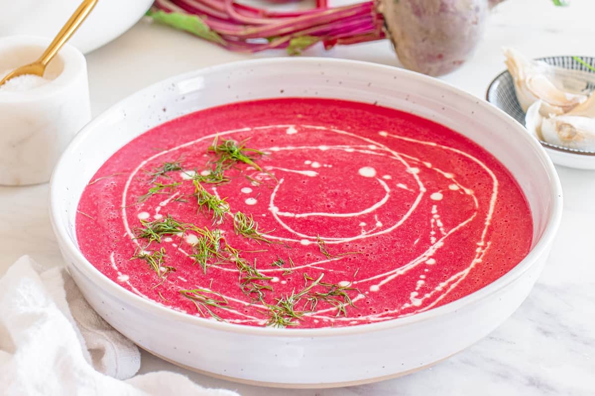 Side angle view of a bowl of beet soup garnished with half and half and herbs.