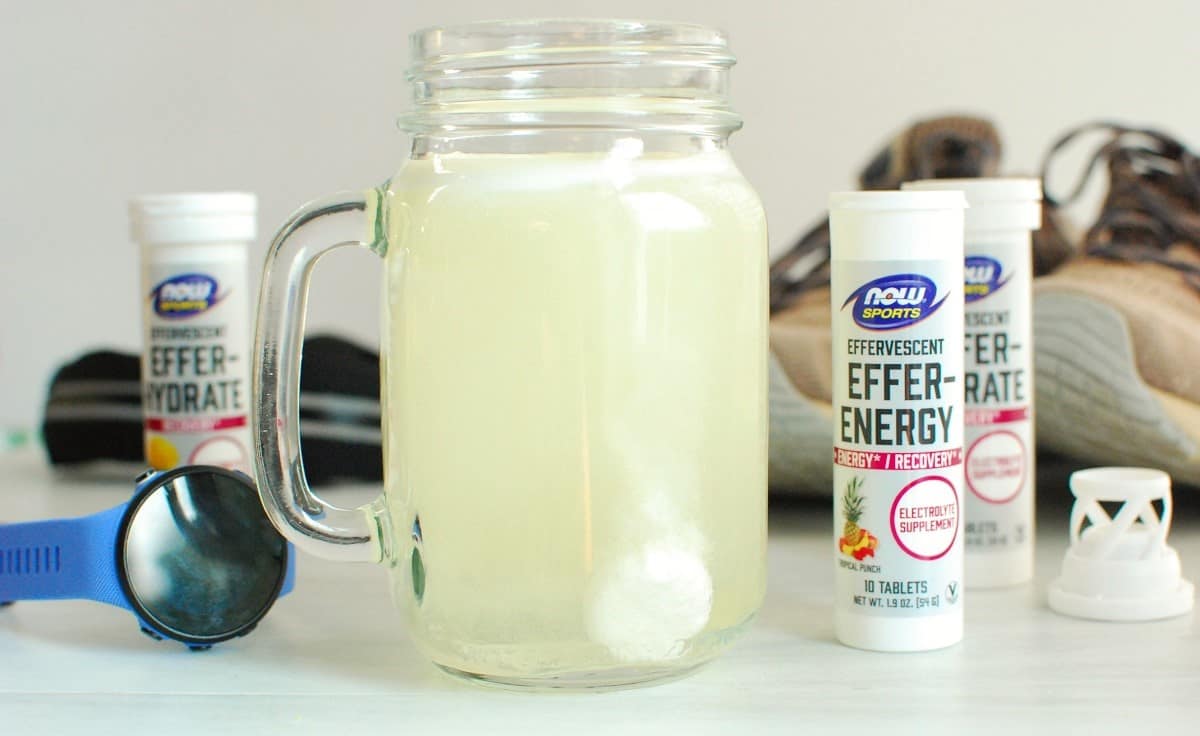 An electrolyte fizz tab dissolving in water in a drinking glass, next to a running watch and sneakers.