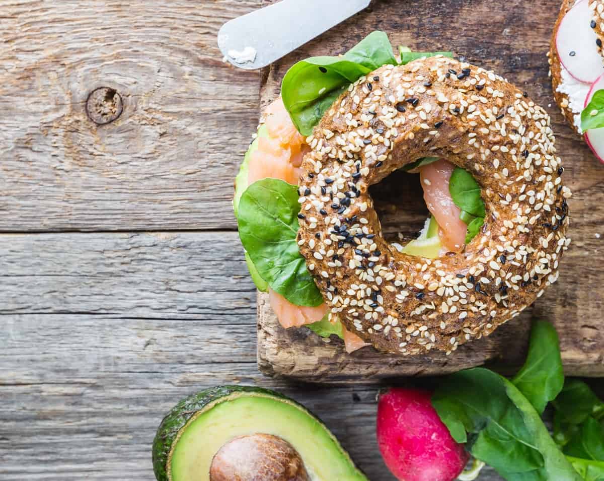 A high protein smoked salmon bagel sandwich on a wooden table next to an avocado.