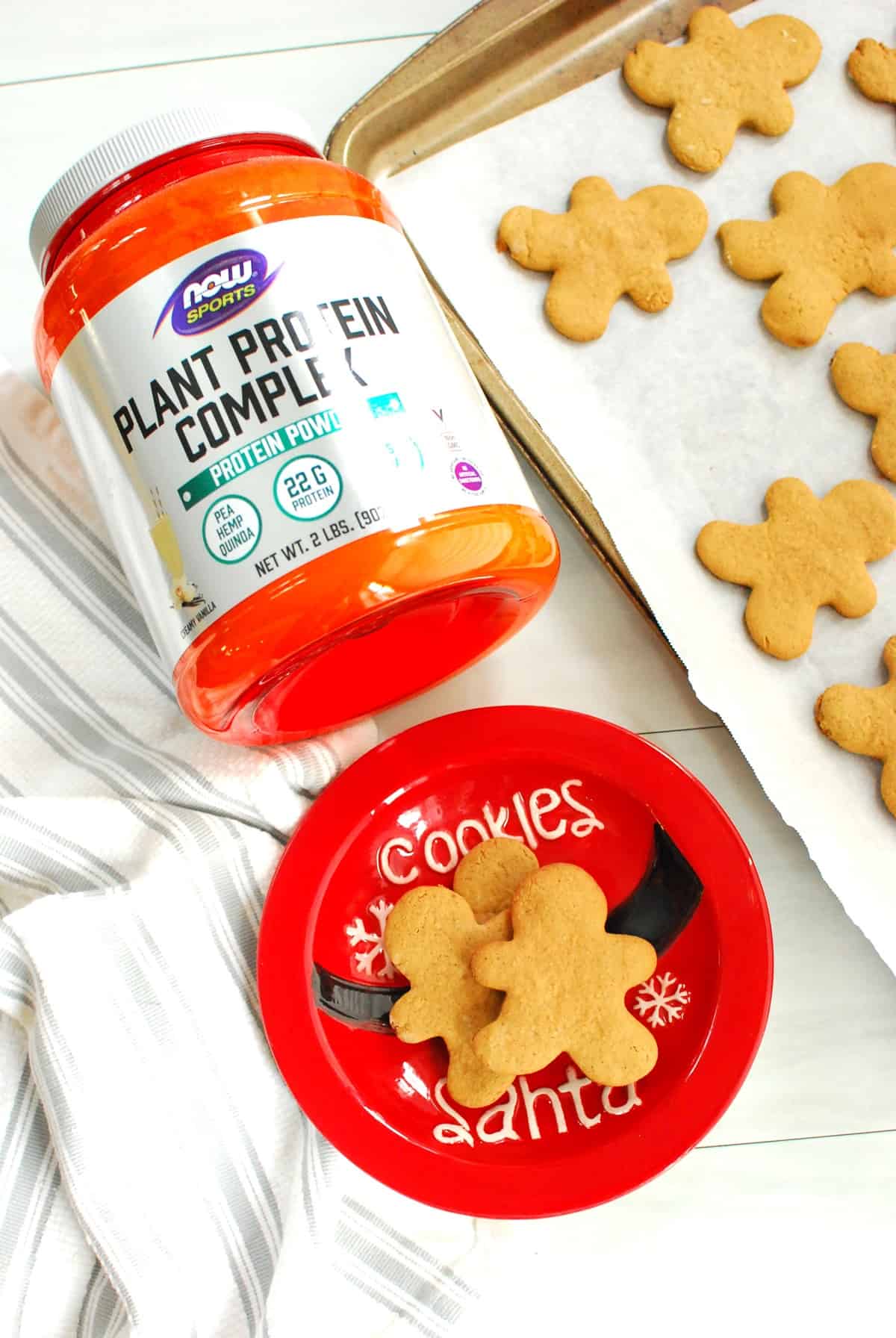 Protein gingerbread cookies on a small red plate next to a container of protein powder, a napkin, and a baking sheet.