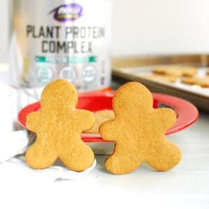 Two protein gingerbread cookies leaning against a plate, with a container of protein powder and a baking sheet in the background.