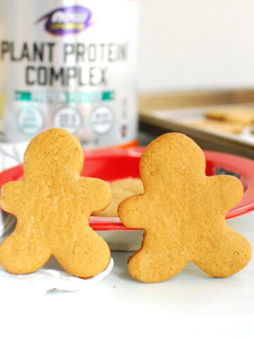 Two protein gingerbread cookies leaning against a plate, with a container of protein powder and a baking sheet in the background.