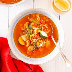 A bowl of chicken vegetable soup with a spoon in it and a red napkin next to the bowl.