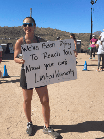 A woman holding a race day sign that says we've been trying to reach you about your car's limited warranty.