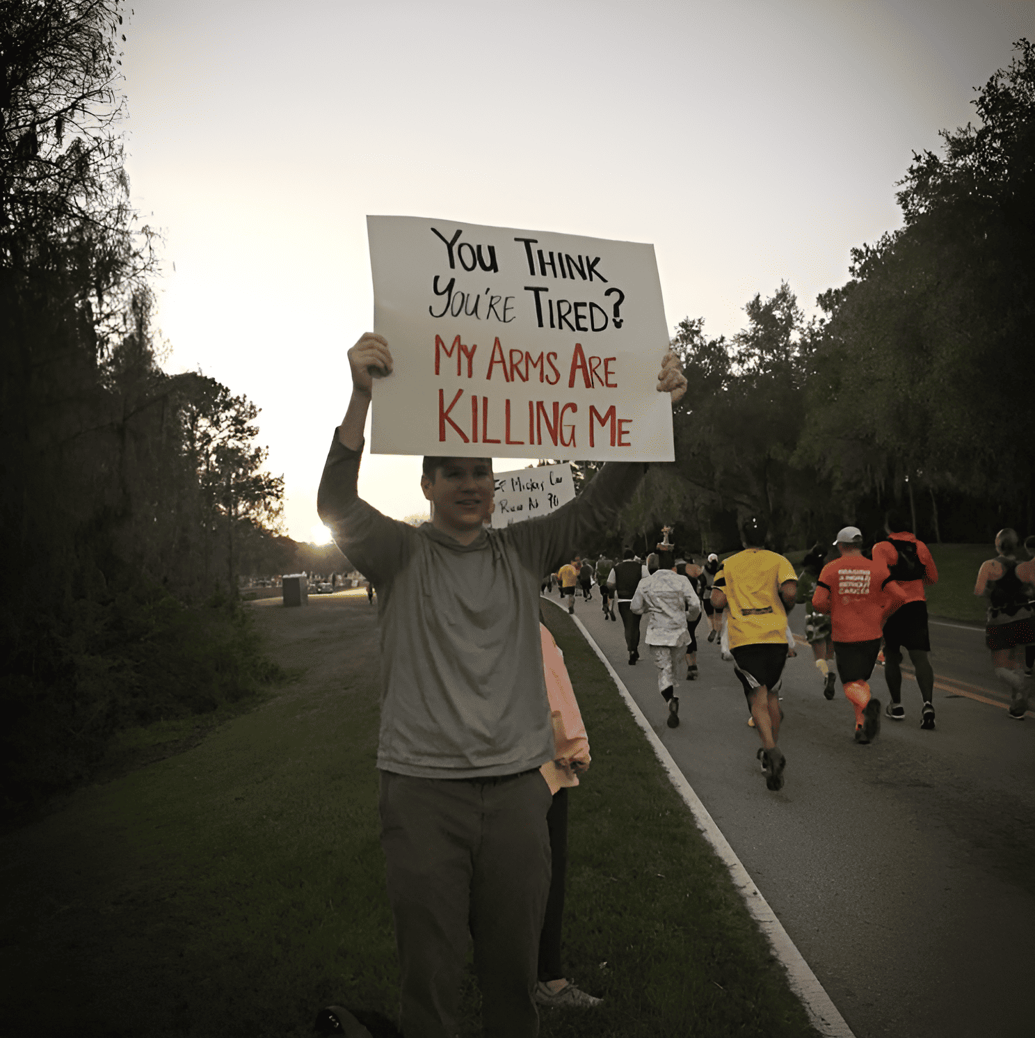 A man holding up a race day sign that says "You think you're tired? My arms are killing me!"