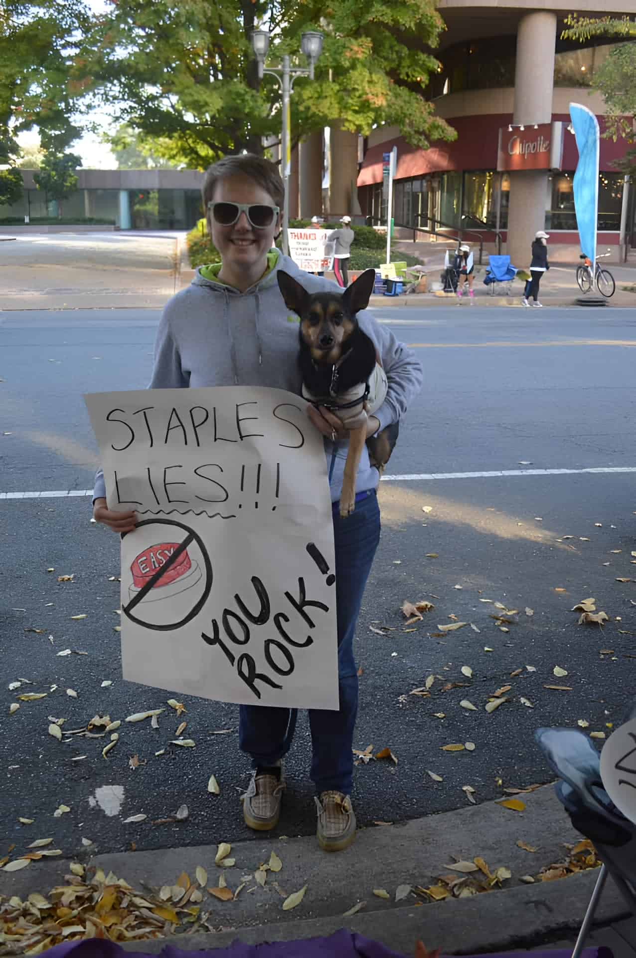 A woman holding a sign at a race with text that says "Staples Lies!" and a crossed out easy button.