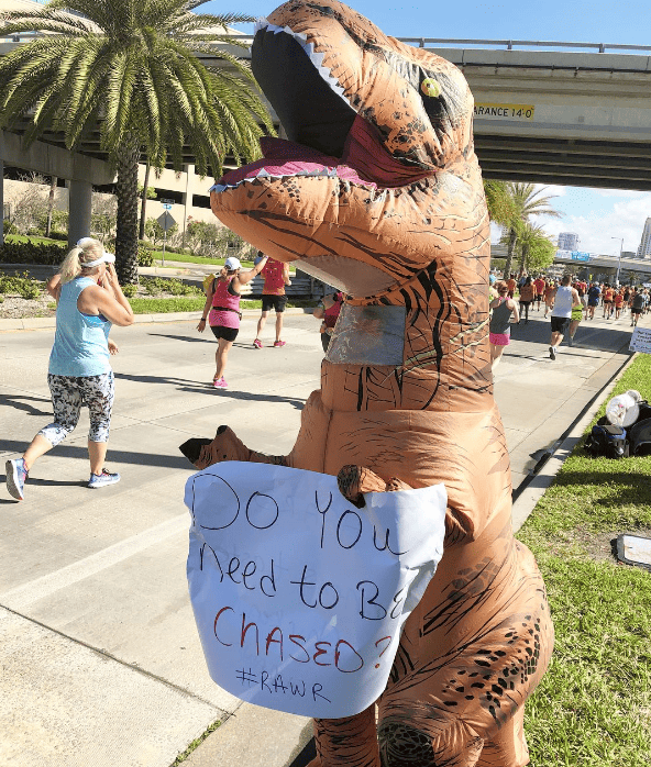 A person in a dinosaur costume holding a sign at a race that says 'do you want to be chased'.