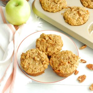 Three healthy morning glory muffins on a plate next to a napkin, apple, nuts, and muffin tin.