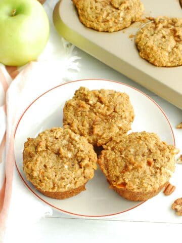 Three healthy morning glory muffins on a plate next to a napkin, apple, nuts, and muffin tin.