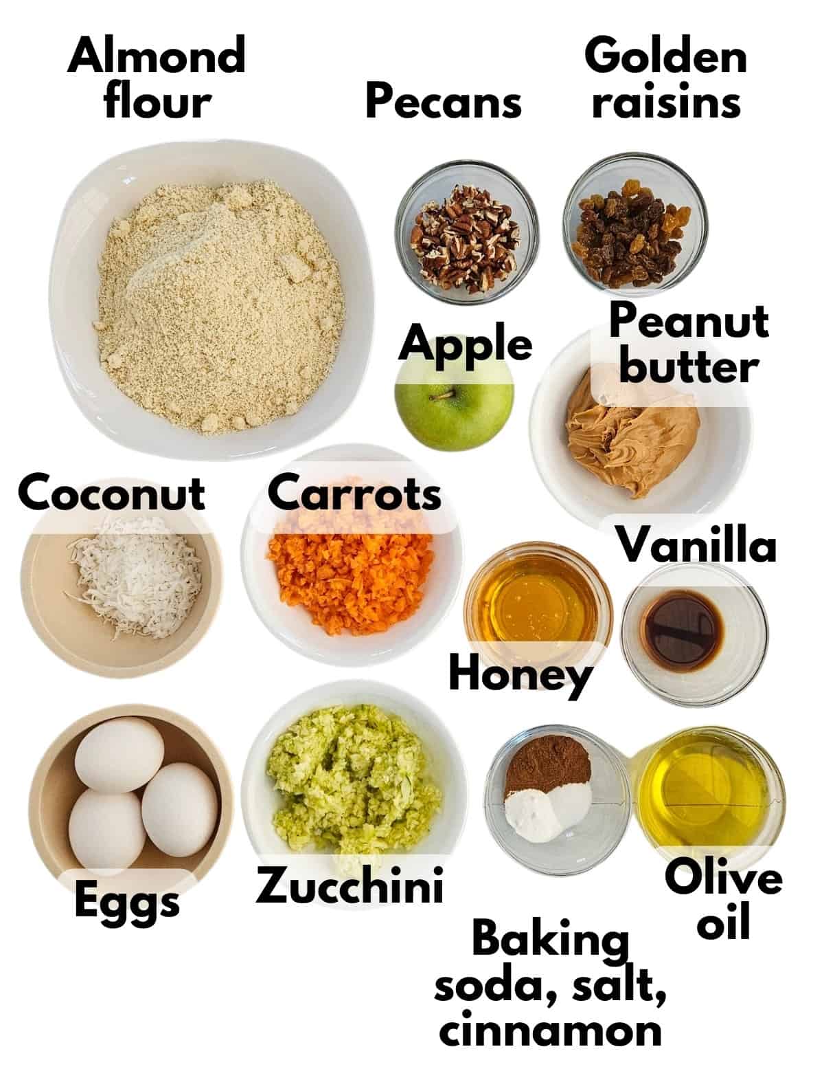 All of the ingredients to make the muffins on a white background, including peanut butter, oil, honey, eggs, vanilla, cinnamon, almond flour, salt, baking soda, apple, carrots, zucchini, coconut raisins, and nuts.